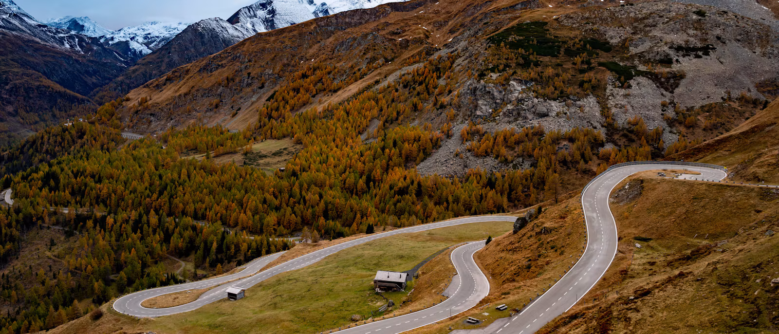 A winding mountain road with hairpin bends in autumn, snow-capped Alpine peaks behind
