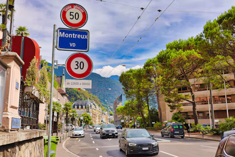 Round speed limit signs on a pole above a street lined with trees and cars, in Switzerland